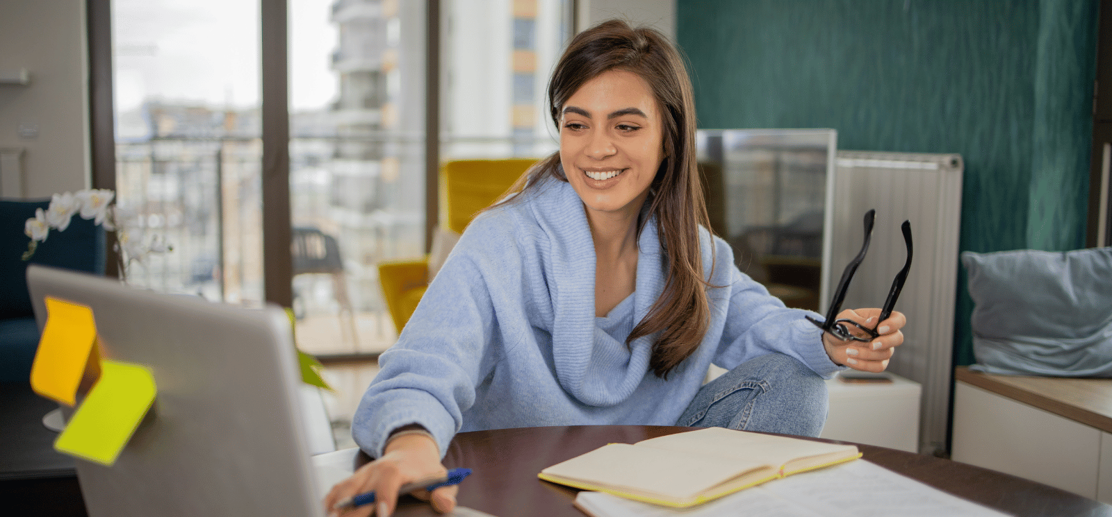 Smiling woman holding eye glasses and sitting at a desk with a laptop.