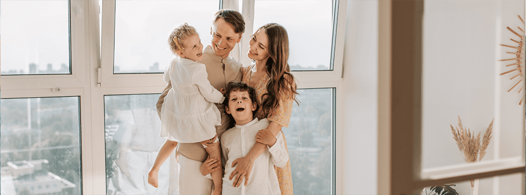 Family of four smiling and standing in front of a window