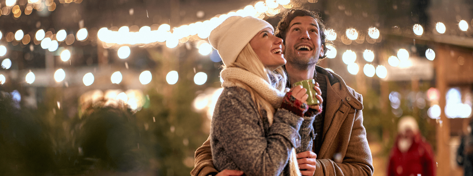 Smiling couple wearing a knit hat and scarf and standing under holiday lights.
