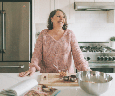 Woman smiling and standing in kitchen with a cookbook and cutting board