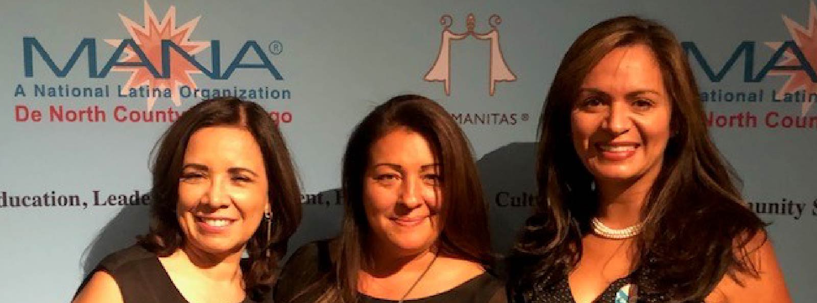 Three women are smiling and standing in front of an event sign.