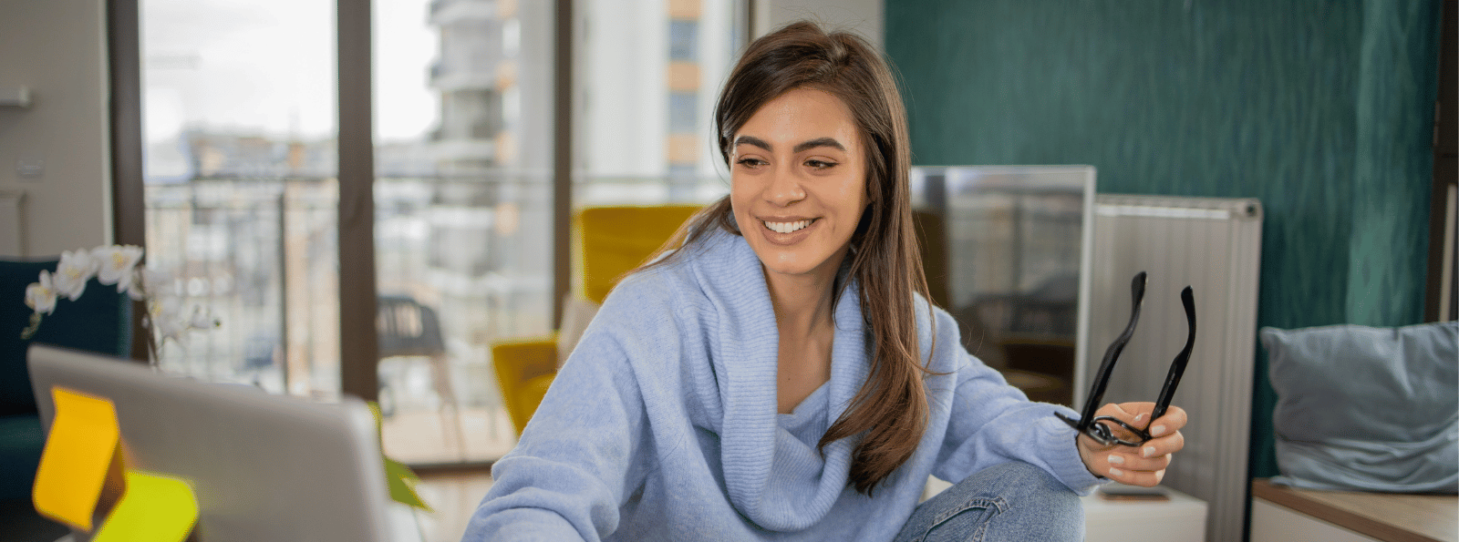 Smiling woman sitting at the computer with eyeglasses in her hand.