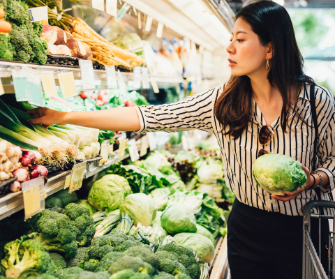 Woman in the produce aisle of a grocery store and has a head of lettuce in her hand.