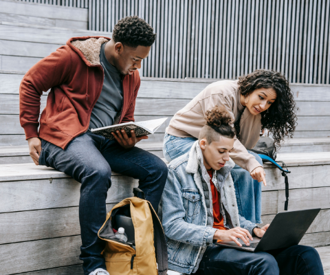 Three adult students sitting on a bench reading a book and looking at a laptop.