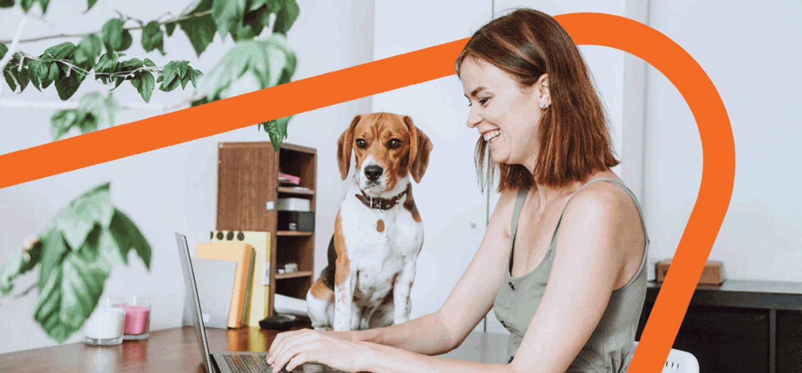 Woman smiling and sitting in a chair at a table working on a laptop computer with a dog sitting on the table looking on.