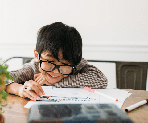 Young boy wearing glasses and sitting at the table working with markers